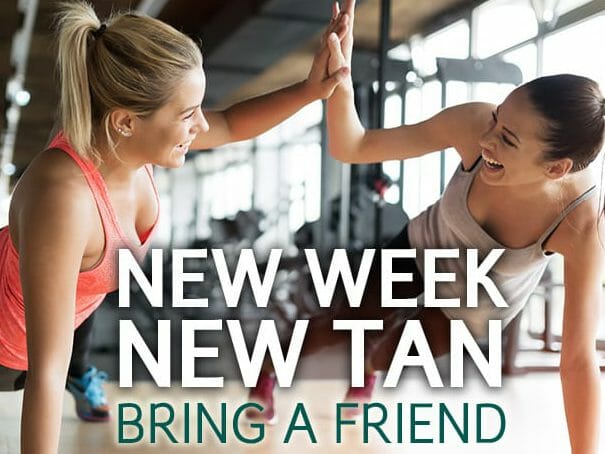 Two women in workout attire high-five each other at a gym. Both are smiling, and the background shows gym equipment and large windows. The text on the image reads, "New Week New Tan, Bring a Friend." Find the best spray tan in New York to keep your glow after every gym session!