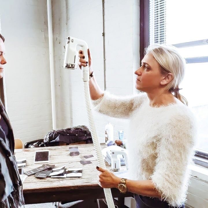 A woman with light hair holds a handheld spray tanning device aimed at another woman's face, who stands still with her eyes closed. They are in an indoor room with large windows, a wooden table, and various items on it.