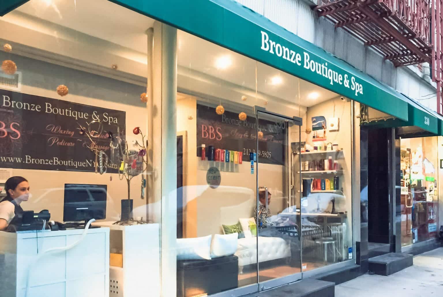 Glass storefront exterior of a spray tanning salon in New york city with green awning with words above it Bronze Boutique & Spa. 