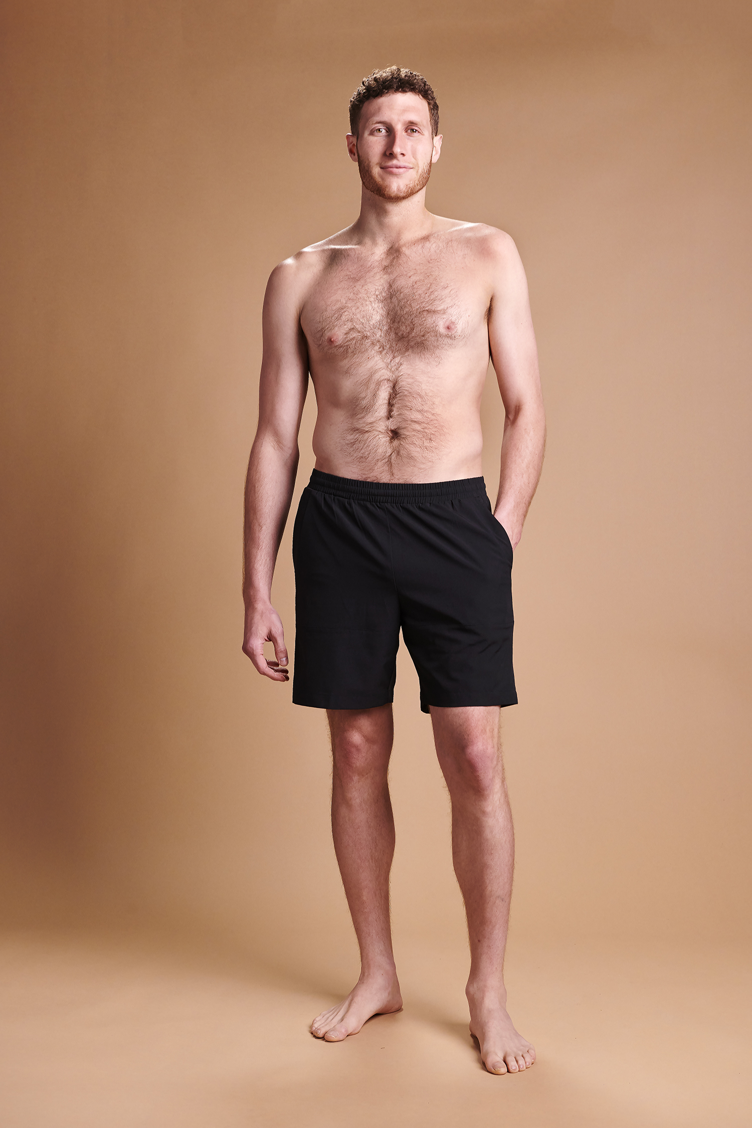 VersaSpa real life light tan picture. A tall, barefoot man with short hair, wearing black athletic shorts, stands against a light brown background. He has his left hand in his pocket while his right arm hangs by his side. He appears relaxed and is looking straight ahead.